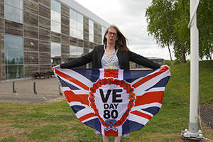 lady, standing in front of a building, holding out teh VE Day 80 flag