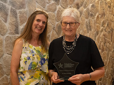 Two ladies standing on a stage. One lady holding an award. Both smiling at the camera.