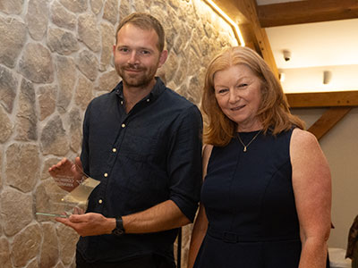Man and lady standing on a stage. Lady holding an award. Both smiling at the camera.