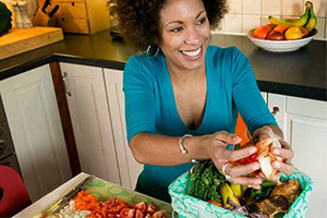 A woman in her kitchen sat at the table with fresh vegetables in a box and on a chopping board