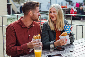 Young man and woman sitting at a bench eating a pasty and having a drink