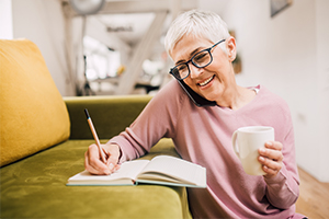 Older lady sat next to a sofa writing on the phone with a mug in one hand and a pen in the other writing in a note pad