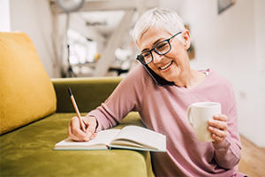 Older lady sat next to a sofa writing on the phone with a mug in one hand and a pen in the other writing in a note pad