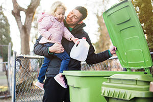 Man holding his young daughter putting a milk carton into a green recycling bin