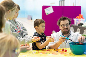 Children stood round a table, mixing food in a bowl with an adult helping them.