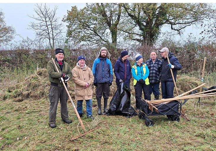 Group photo of people standing in a meadow holding various gardening tools, smiling at the camera