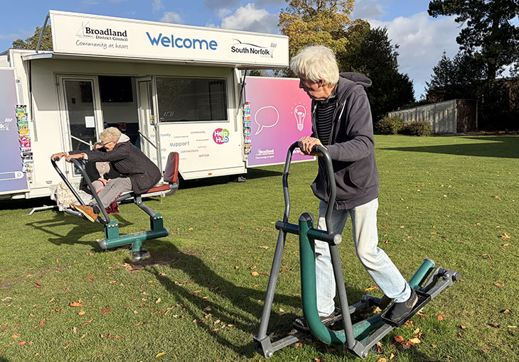 Help Hub van with doors open on a playing field. A lady and a man on the outdoor gym equipment, in front of the van