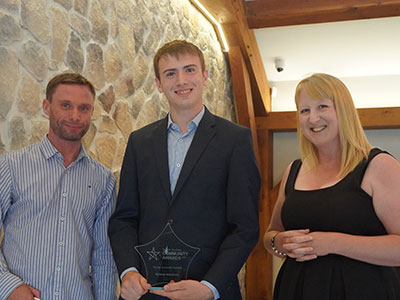 Young man, standing on a stage holding an award, with a lady and man on each side of him.