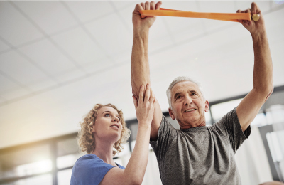 An older man doing stretches with an exercise band above his head and a woman assisting him