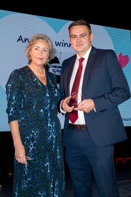 Lady in a green dress with short grey hair, presenting an award to a man in a blue suit with a red tie