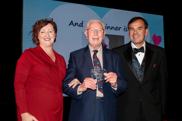 Lady in a red dress with red hair and a man in a suit with a bow tie presenting an award to an older man in a blue suit