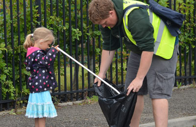 a young girl putting litter into a bin bag being held by a man wearing a high vis jacket