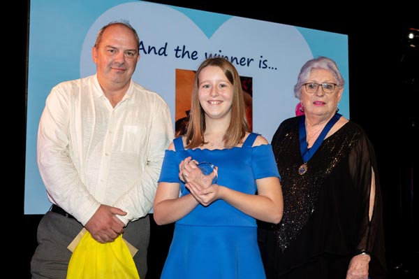 A man in a white shirt and grey trousers with a lady in a black dress and vice chairman chain present an award to a young girl in blue dress